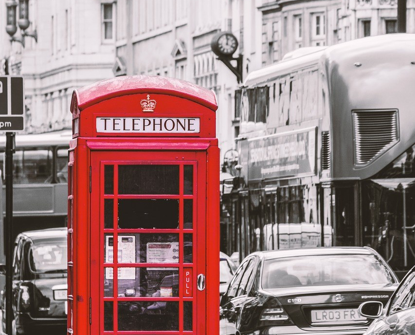IMAGE DESCRIPTION: A bright red British telephone booth sits in the foreground of a black and white photograph of a busy London street filled with cars and double-decker busses.